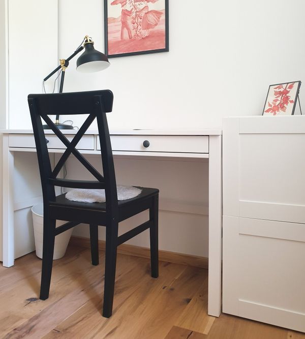 Ein weißer Schreibtisch mit schwarzem Stuhl und Schreibtischlampe. In unseren Zimmern stellen wir Ihnen auch einen Arbeitsplatz zur Verfügung.